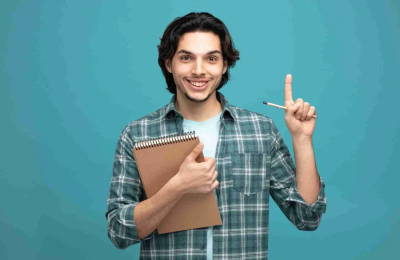 A smiling young man in a plaid shirt holds a notepad and pen, giving a thumbs-up and raising his index finger, ready to share techniques for preparing for IELTS against a solid teal background.
