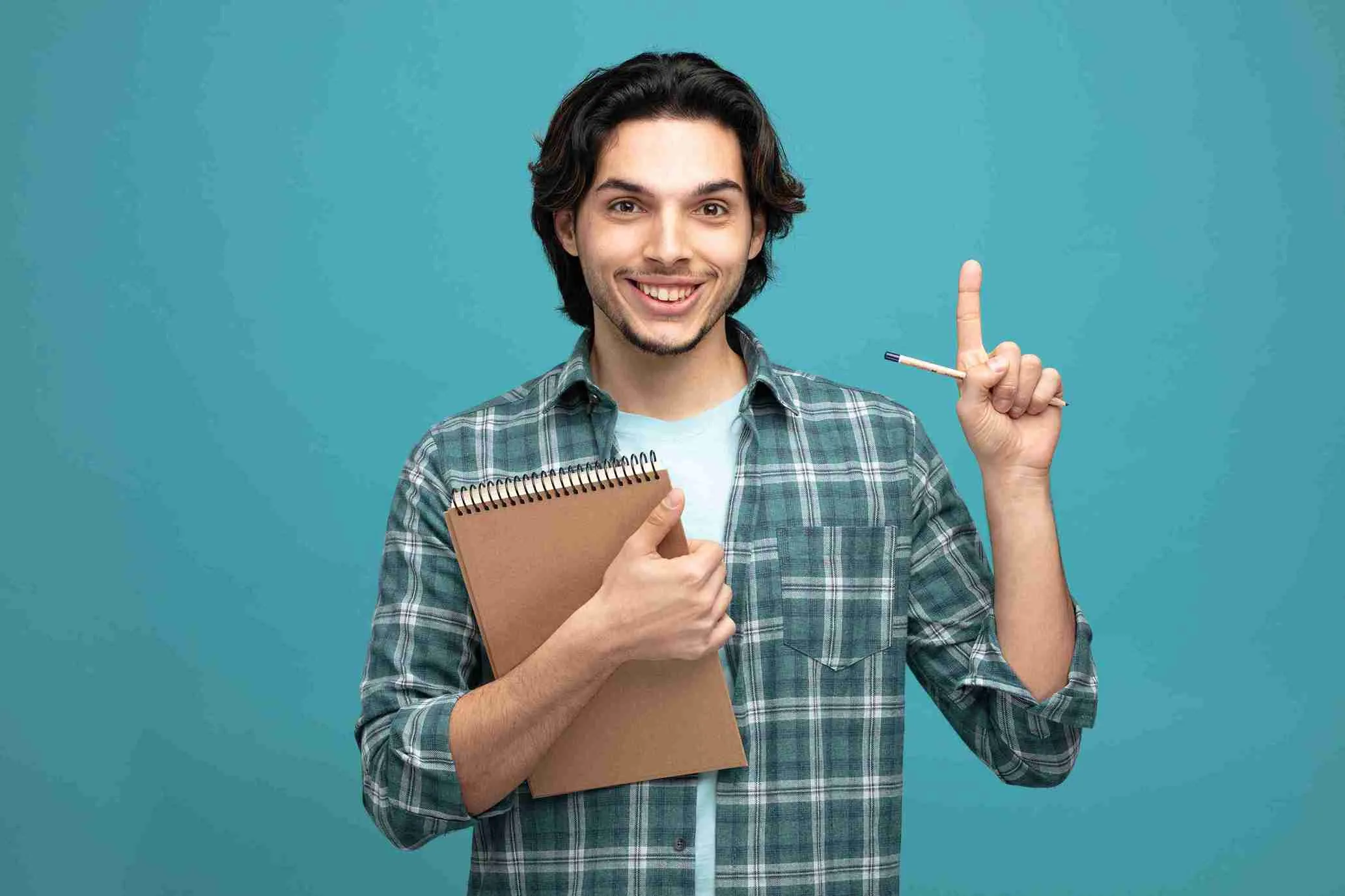 A smiling young man in a plaid shirt holds a notepad and pen, giving a thumbs-up and raising his index finger, ready to share techniques for preparing for IELTS against a solid teal background.