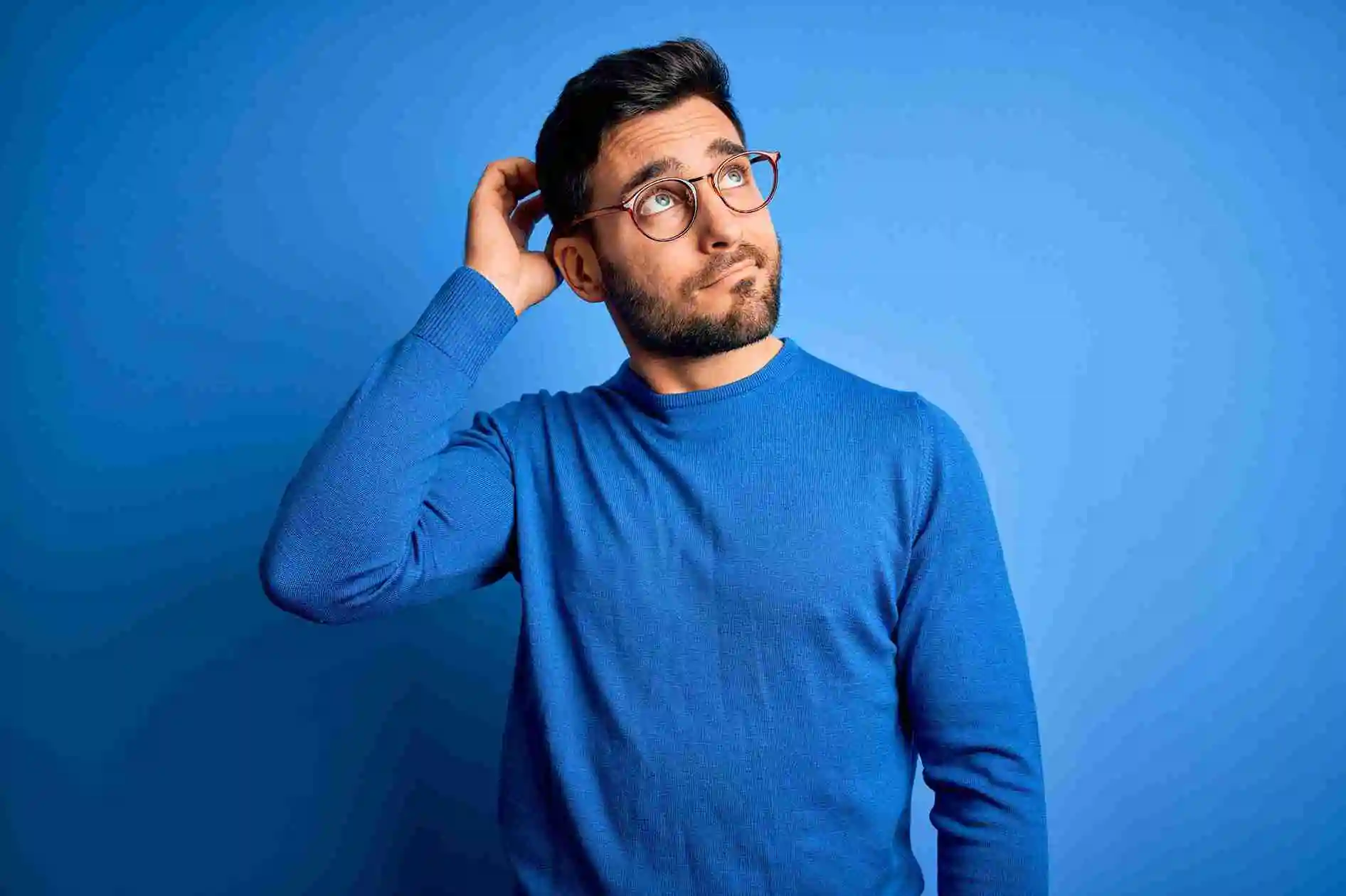 A man with dark hair, a beard, and glasses wearing a blue sweater stands against a blue background, scratching his head and looking upward with a thoughtful expression as if wondering, Can we clear IELTS without coaching?.