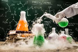 A scientist in a lab coat pours a green liquid from a flask into a beaker, surrounded by colorful chemical flasks and a chalkboard with formulas.