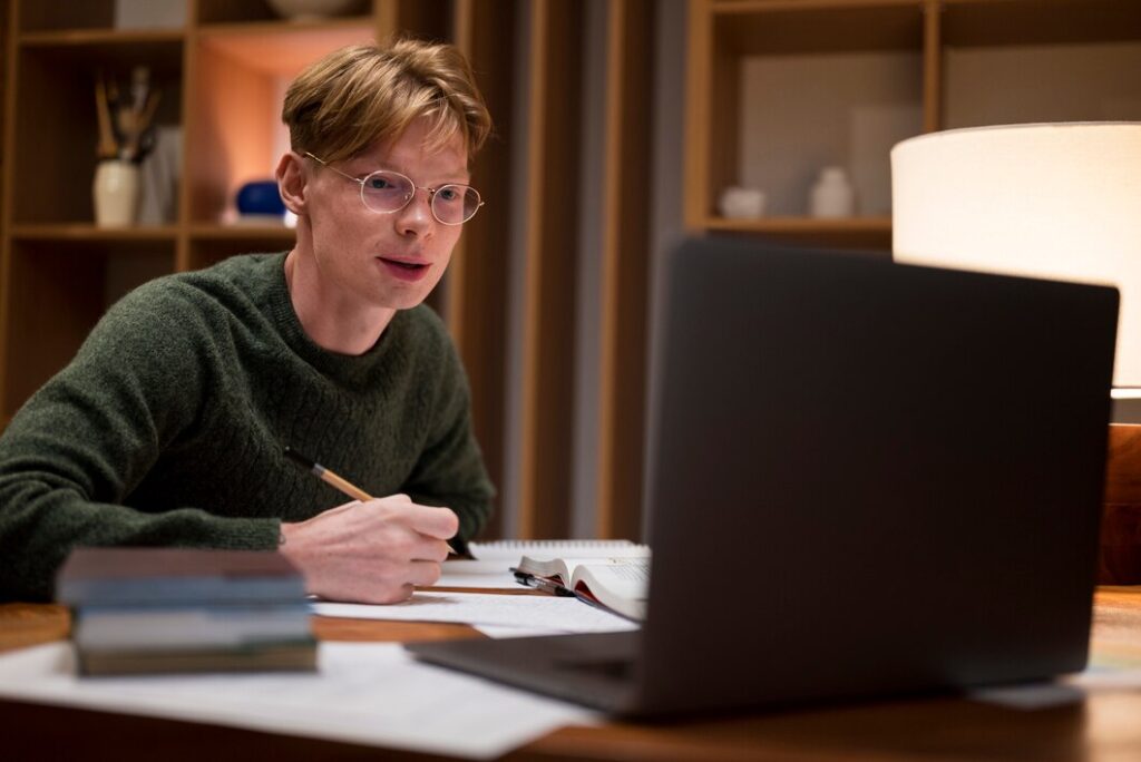 A person in a green sweater writes notes at a desk, with a laptop, books, and papers in a well-lit room.