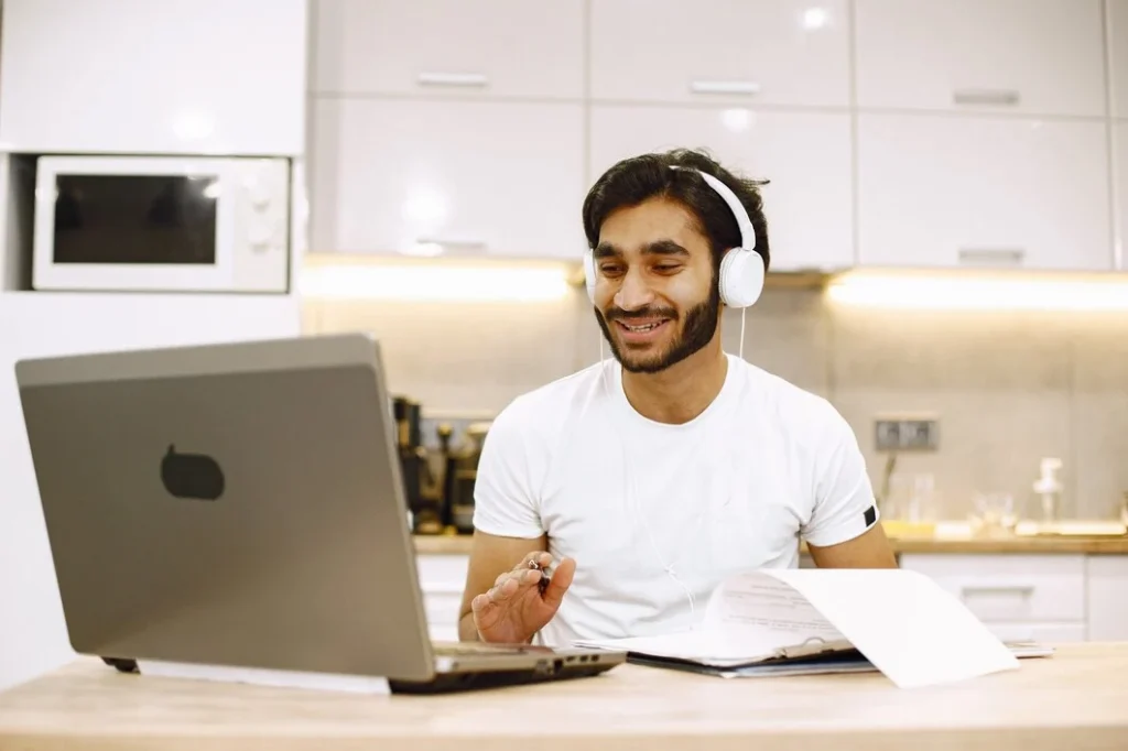 A person wearing headphones sits at a wooden table, using a laptop with papers and books in front of them, in a bright kitchen.