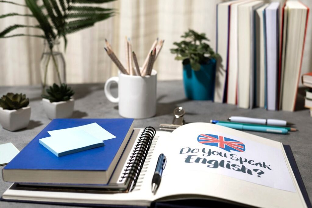 A study desk featuring a notebook open with "Do You Speak English?" text, surrounded by books, stationery, and plants.