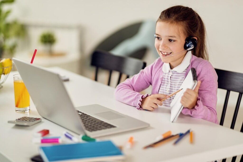 A young girl sits at a table, focused on her laptop while wearing headphones.