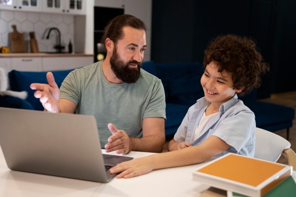 A bearded man and a smiling boy interact with a laptop at a table. The scene is warm and inviting, set in a modern kitchen with a cozy atmosphere.